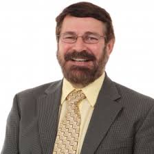 Dr. Randy Brooks, who has short brown hair and wire glasses. He is wearing a grey suit with a yellow shirt and yellow, printed tie.