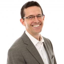 Dr. Stephen Frech, who has short brown hair and black glasses. He has black hair and is wearing a brown suit with a white button-up shirt.